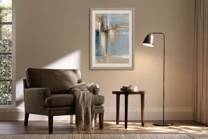 Framed abstract artwork above a dark upholstered armchair beside a floor lamp and small table.