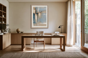 Framed abstract artwork above a wooden desk with a chair, lamp, and open glass door.