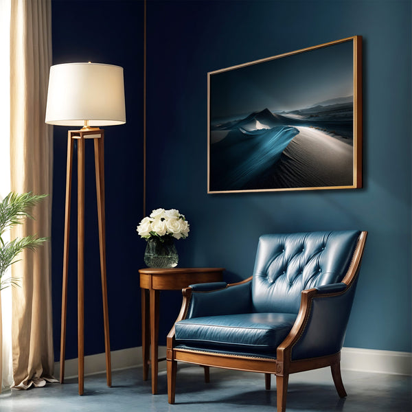 Blue-toned desert dune artwork in a thin gold frame above a blue leather armchair.