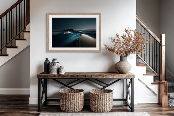 Matted desert dune print in a light wood frame above a console table between staircases.