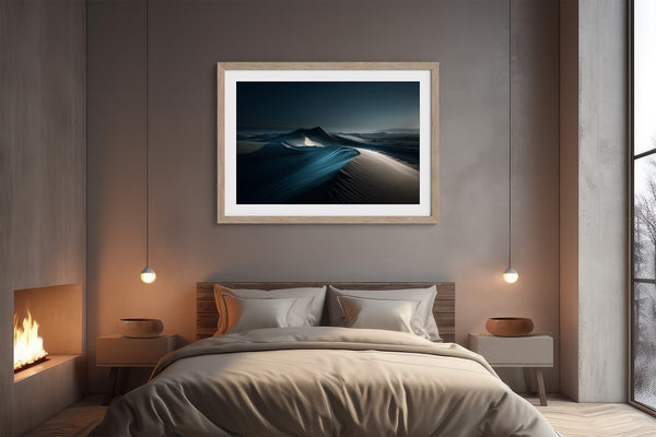 Matted desert dune artwork in a light wood frame centered above a bed.