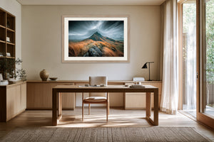 Framed stormy mountain ridge artwork displayed above a wooden desk beside tall windows.