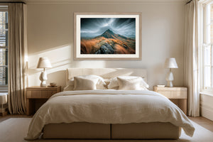Framed stormy mountain artwork centered above a bed with cream bedding and bedside lamps.