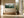 Framed misty pine forest watercolor above a built-in bench beside tall black-framed windows.
