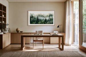 Framed misty pine forest watercolor above a wooden desk with a chair and black task lamp.