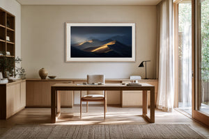 White-framed dune artwork above a wooden desk beside tall windows and sheer curtains.