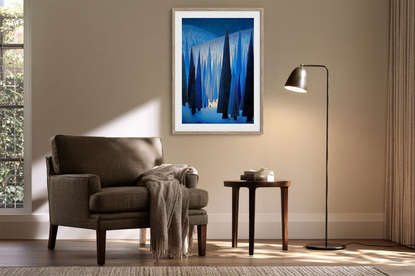 White-framed blue conifer artwork above an upholstered armchair, side table, and floor lamp.