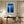 White-framed blue forest print above a wooden desk and swivel chair by sheer curtains.