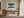 Framed Leaning Light tree artwork above a wooden desk near tall windows and sheer curtains.