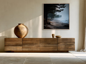 Framed misty pine shoreline artwork above a wood sideboard with ceramic vessels.