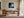 Framed misty pine shoreline artwork above a wooden desk near tall windows.