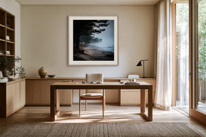 Framed misty pine shoreline artwork above a wooden desk near tall windows.