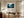 Light wood-framed misty conifer forest artwork above a wooden desk and swivel chair.
