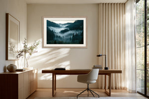Light wood-framed misty conifer forest artwork above a wooden desk and swivel chair.