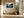 Light wood-framed misty forest artwork above a built-in bench beside large black-framed windows.