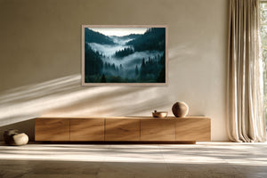 Light wood-framed misty conifer forest artwork above a long wooden sideboard in a sunlit space.