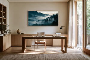Framed misty forest artwork above a wooden desk beside tall windows.