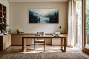 Framed misty conifer lake artwork above a wooden desk beside tall windows.