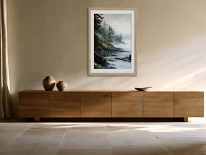 Misty evergreen shoreline artwork in a pale wood frame above a long wooden sideboard.