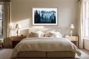 Light wood-framed misty pine forest artwork above a bed with cream bedding and matching bedside lamps.