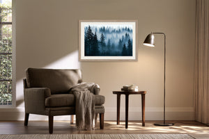 Light wood-framed misty pine forest artwork above a dark armchair, side table, and black floor lamp.