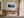 Light wood-framed misty pine forest artwork above a wooden desk in a bright workspace.