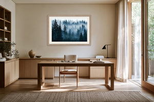 Light wood-framed misty pine forest artwork above a wooden desk in a bright workspace.