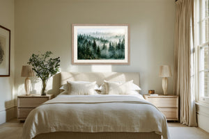 Light wood-framed misty evergreen forest artwork above a white bed between two bedside tables.