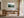 Light wood-framed evergreen forest artwork above a wooden desk and chair beside tall windows.