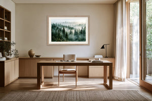 Light wood-framed evergreen forest artwork above a wooden desk and chair beside tall windows.