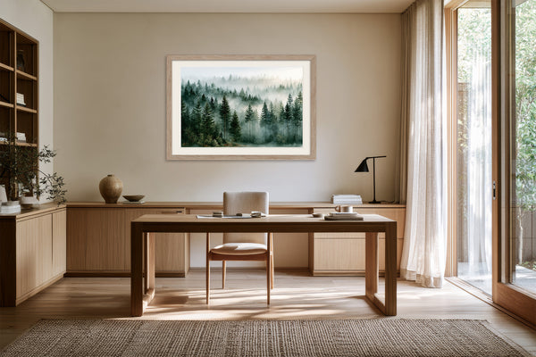 Light wood-framed evergreen forest artwork above a wooden desk and chair beside tall windows.