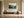 Light wood-framed misty forest artwork above a low wooden bench beside a large ceramic vessel.