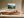 Light wood-framed evergreen forest artwork above a long wooden cabinet in a sunlit neutral interior.