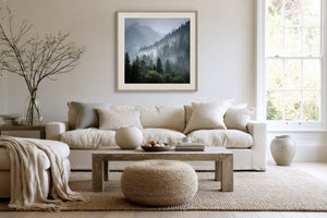 Framed misty mountain forest artwork above a cream sofa, with a wood coffee table and round ottoman.