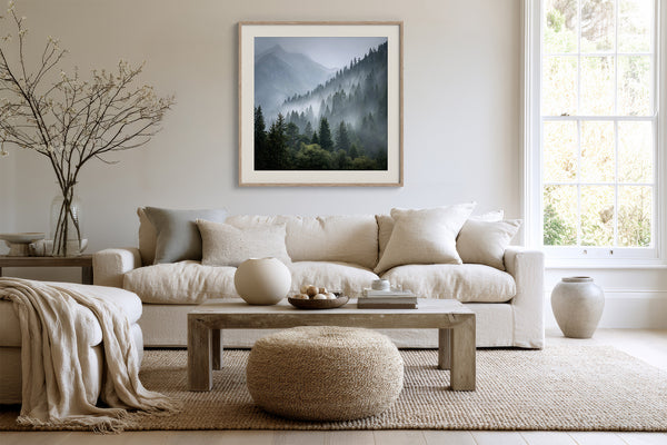 Framed misty mountain forest artwork above a cream sofa, with a wood coffee table and round ottoman.