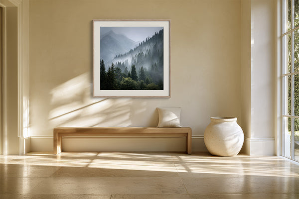 White-framed misty mountain forest artwork above a wooden bench beside a large ceramic vase.