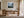 White-framed misty mountain forest artwork above a wooden desk with chair and black task lamp.