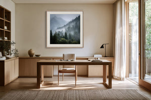 White-framed misty mountain forest artwork above a wooden desk with chair and black task lamp.