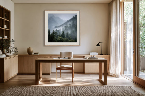 White-framed misty mountain forest artwork above a wooden desk with chair and black task lamp.