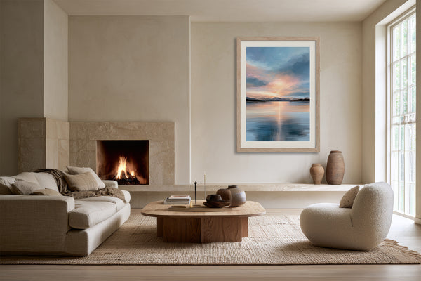 Light wood-framed mountain lake artwork above a low console near a fireplace and curved chair.
