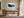 Framed snowy pine forest artwork above a wooden desk beside tall glass doors.