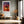 Light wood-framed sunset tree print above a bench beside tall windows.
