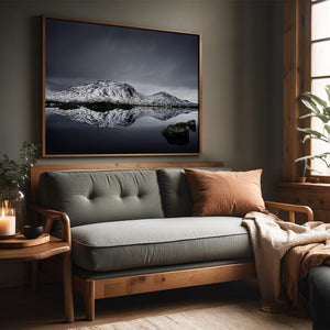 Large dark-framed snowy mountain reflection print above a gray sofa beside a wood-trimmed window.