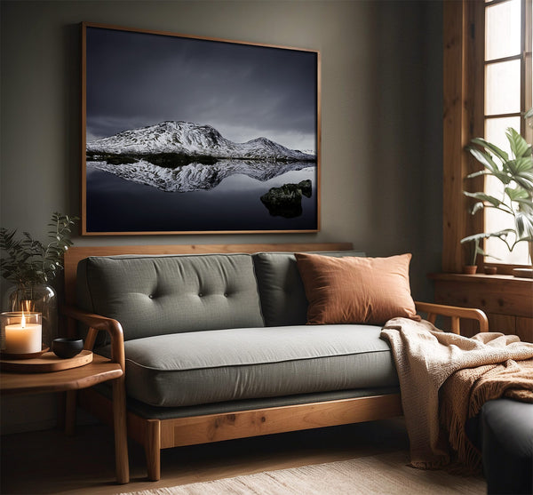 Large dark-framed snowy mountain reflection print above a gray sofa beside a wood-trimmed window.