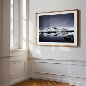 Light wood-framed mountain reflection print displayed on a white paneled wall beside tall windows.
