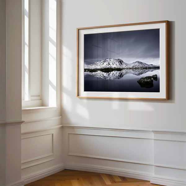 Light wood-framed mountain reflection print displayed on a white paneled wall beside tall windows.