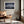 Light wood-framed mountain reflection print above a tan leather chair near a sunlit window.
