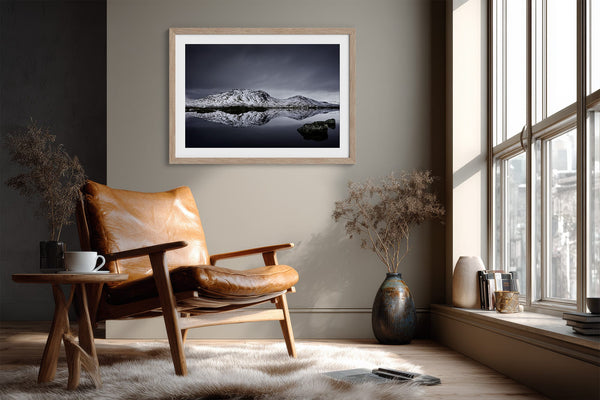 Light wood-framed mountain reflection print above a tan leather chair near a sunlit window.