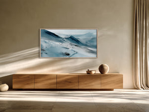 Framed snowy mountain landscape above a long wooden sideboard beside tall beige curtains.