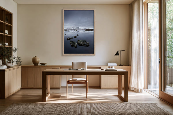 Light wood-framed mountain lake artwork above a wooden desk beside tall windows.